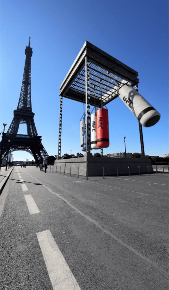 Large UFC punching bags hanging from a structure in an open plaza near the Eiffel Tower